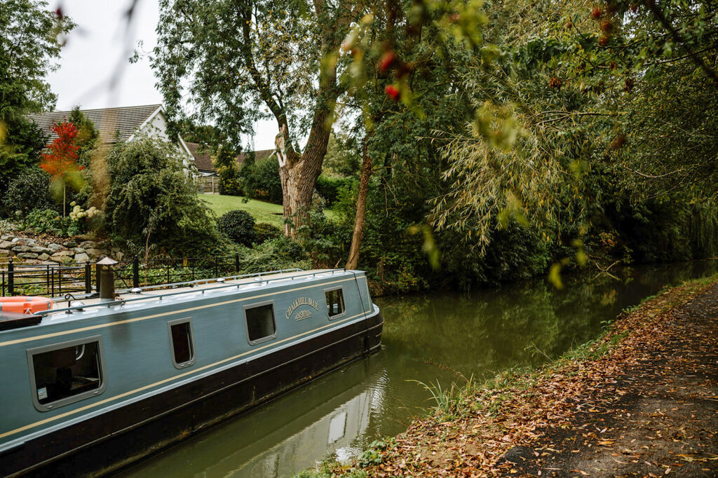 Luxury Boutique Narrowboat moored on a UK canal in autumn, surrounded by golden foliage and calm reflective waters, perfect for a tranquil narrowboat holiday