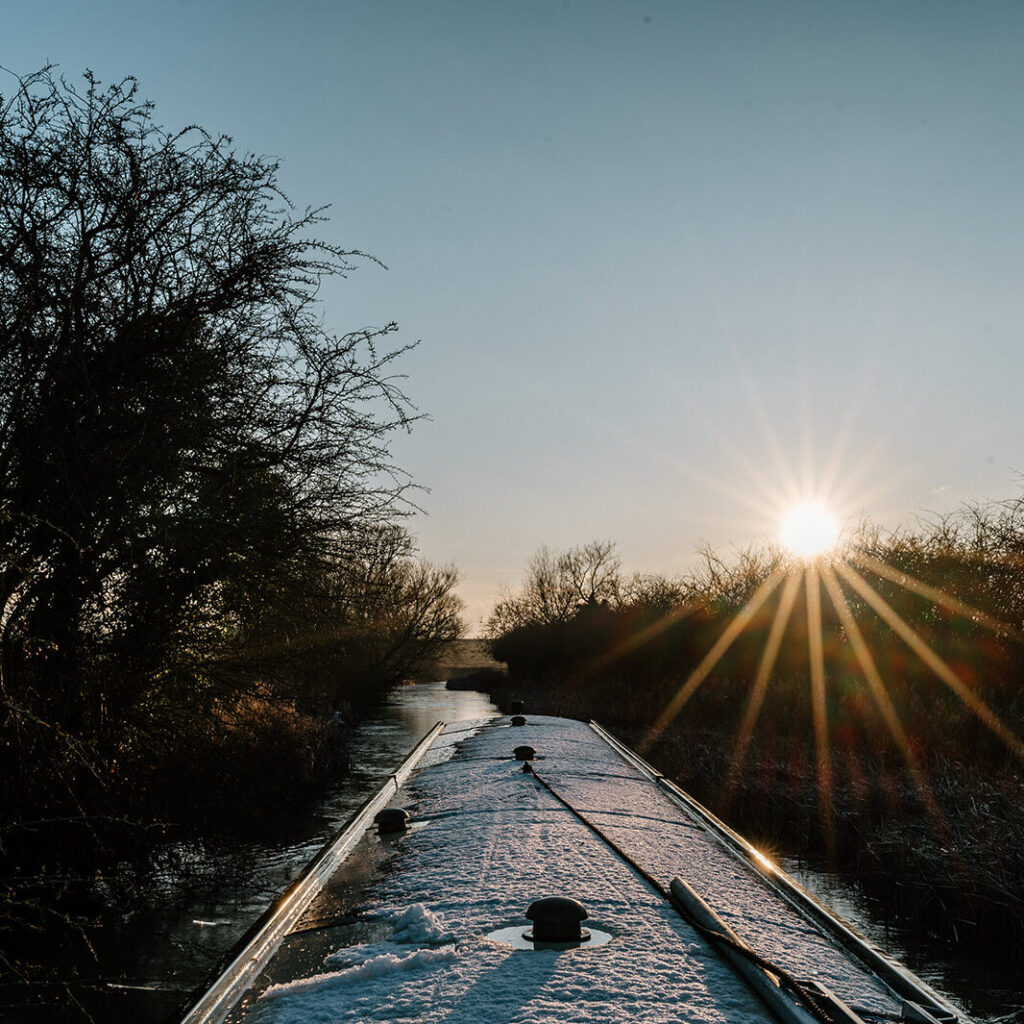Narrowboat cruising along a winter canal at sunset, with soft pink skies ahead and snow settled on the roof - a peaceful seasonal scene capturing the magic of cold-weather boating.