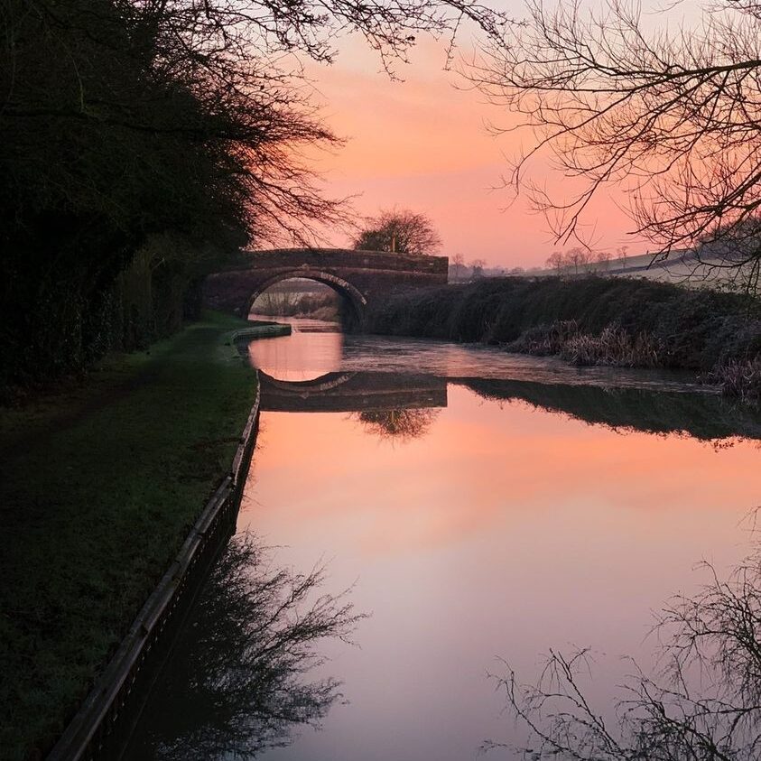 Pink winter sunset reflecting over a calm canal, with bare trees lining the towpath and frost on the ground - a peaceful seasonal scene perfect for a romantic narrowboat holiday.