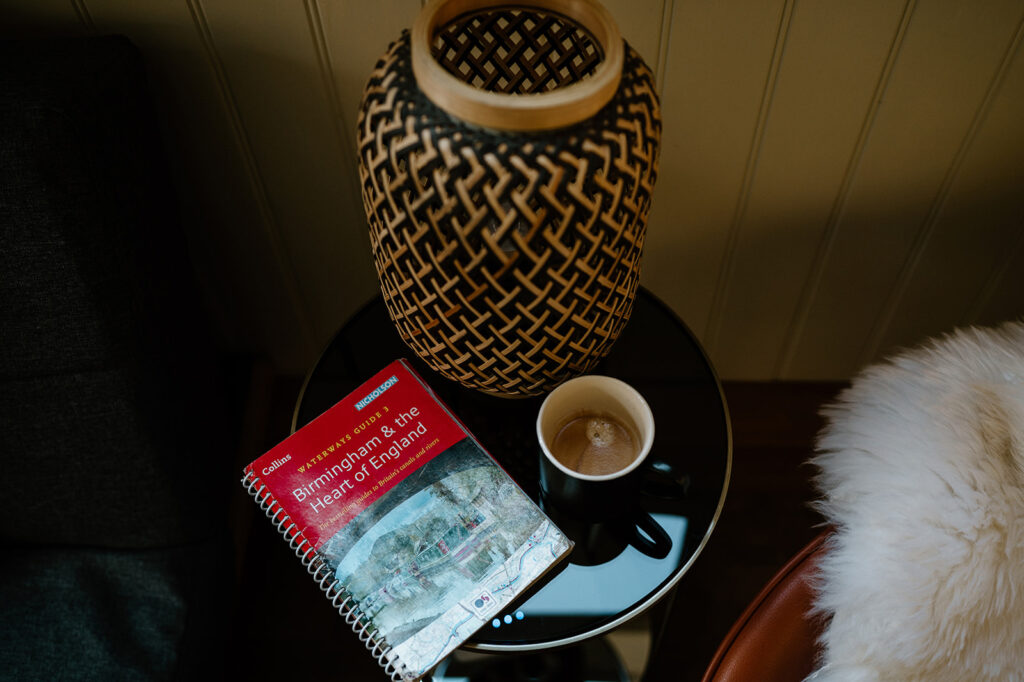 Coffee cup, folded canal map, and a small lamp arranged on a table inside a narrowboat, with soft natural light from a nearby window