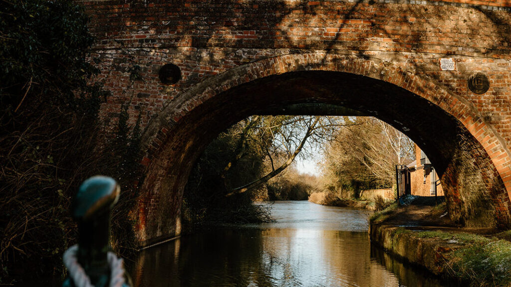 Front of a narrowboat approaching a brick canal bridge, with calm water .