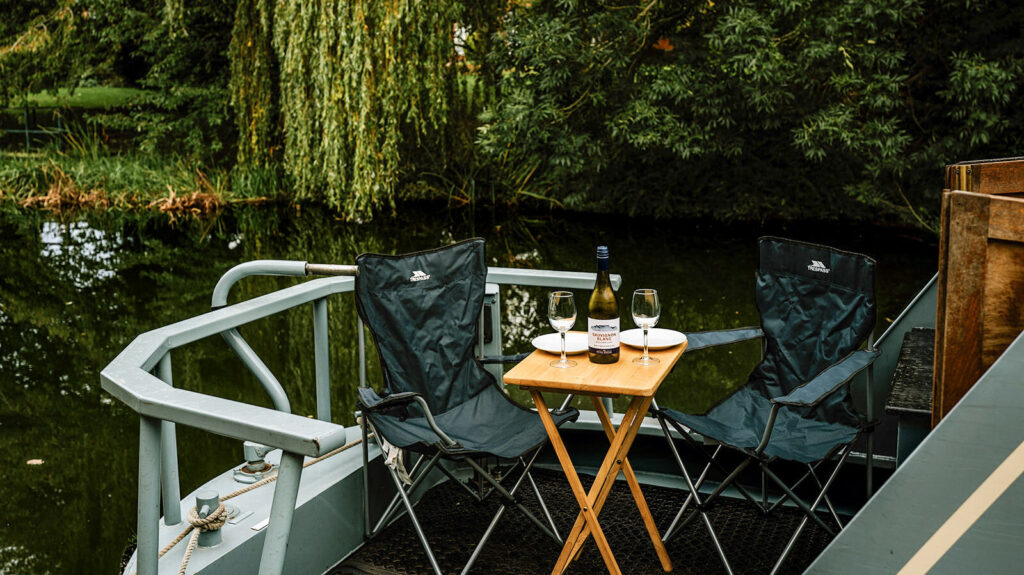 Camp chairs and a small table arranged on the back deck of a canal boat, overlooking a peaceful canal with greenery in the background.