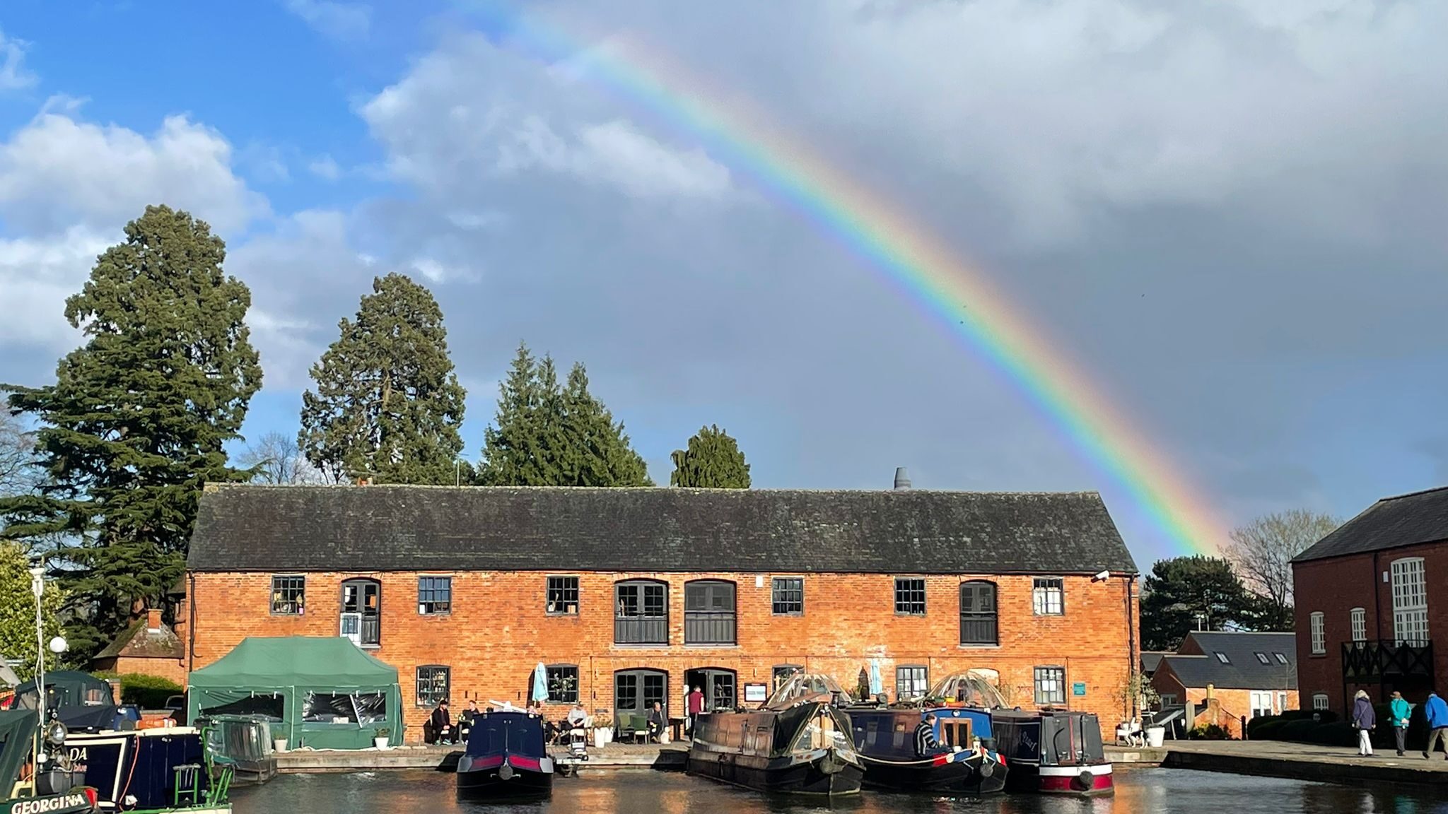 A vibrant rainbow arches over the Waterfront Restaurant, with canal boats moored along the peaceful waterway beneath a partly cloudy sky