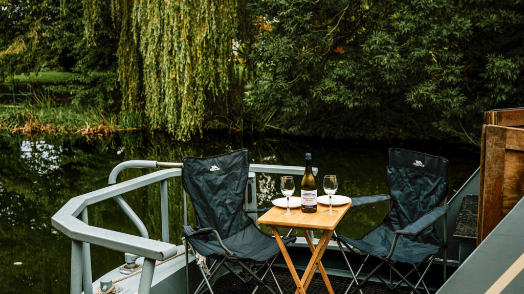 Outdoor seating set up beside a narrowboat, overlooking calm water and greenery.