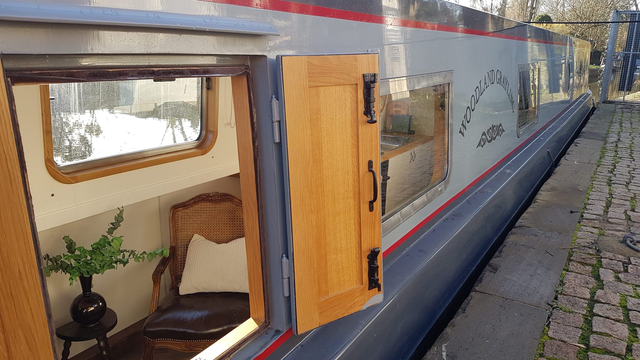 Exterior of a dog friendly narrowboat moored at the waterside, showing cabin windows and wooden doorway beside the canal.