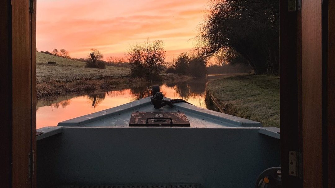 View from the bow of a narrowboat cruising toward a sunset over calm spring water.